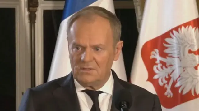 Headshot of Tusk speaking, he wears a suit and stands in front of flags