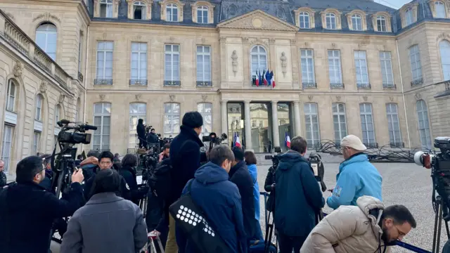 Journalists are gathered with their cameras and equipment in the courtyard of the Elysee Palace. The building is grand and flying French and EU flags