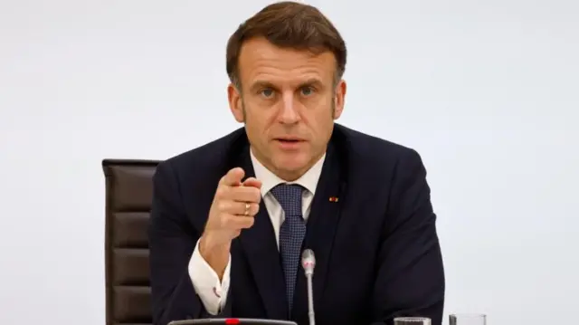 French President Emmanuel Macron sits in a chair wearing a suit, pointing at the camera