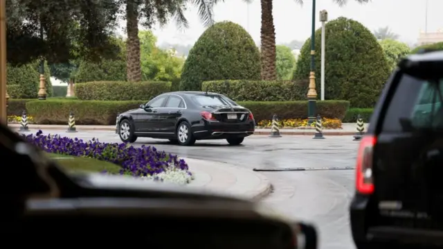 A car driving away on a curved road. There are trees and well trimmed hedges in the background, bollards along one side of the road, and on the left side of the car there's nice lawn and purple flowers bordering the path. Two cars are in the foreground and blurry, you can only see the back of one and the front of the other