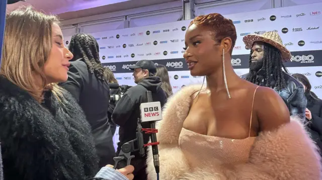 Nigerian singer-songwriter Ayra Starr wearing long earrings and a pale pink dress speaks the BBC on the red carpet