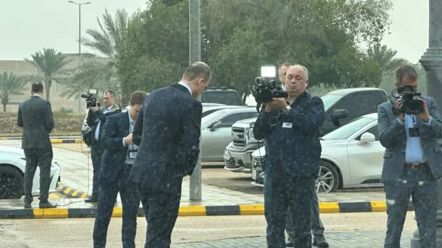 Reporters dressed in suits speak to cameras with cars in the background