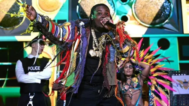 Byron Messia sings into a microphone. He is wearing a colourful jacket, made up of colourful string.