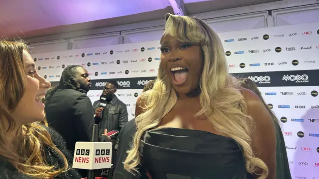 Nella Rose speaks to the BBC on the red carpet at the Mobo awards. She is wearing a black dress and have black hair that fades to blonde. She has brown eyes.