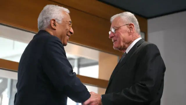 Costa on the left smiles as he shakes hands with Kellogg on the right. Kellogg also holds Costa's arm with his hand. Both wear glasses and have grey hair. They also wear suits.