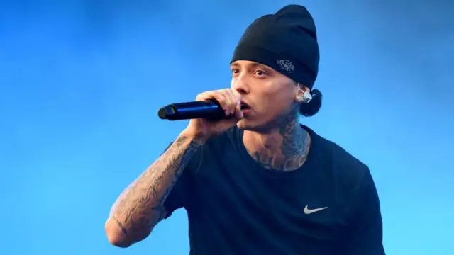 Central Cee sings into a microphone. He is wearing a black top and a black beanie. He has tattoos on his arm and neck.