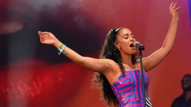 Jorja Smith is singing into a microphone. She is wearing a blue and red striped top.