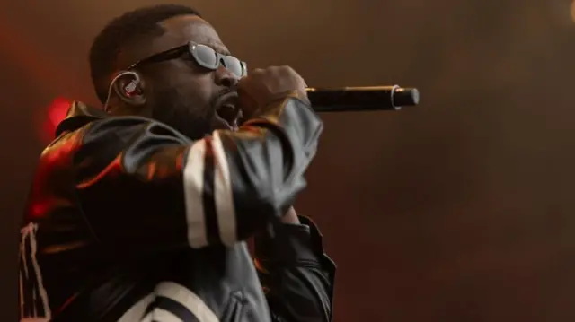 Ghetts sings into a microphone. He is wearing a black leather jacket and black sunglasses.