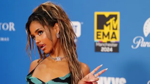 Tyla poses for the camera, doing a peace sign with her left hand and sticking her tongue out the side of her mouth. She is wearing a blue and black striped dress and a silver necklace and earrings.