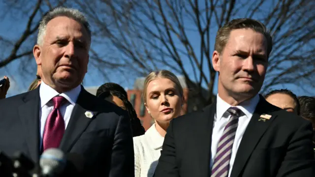 Two men in suits stand next to each other. The sun is shining on their faces. In the background are a crowd of people, including a woman in between them.