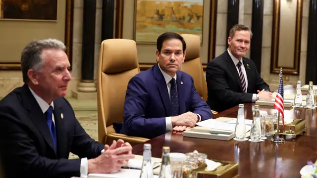 The US delegation sit in front of a US flag at a wooden table