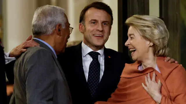 Emmanuel Macron laughs as he embraces Antonio Costa and Ursula von der Leyen. All three are smiling and look happy.