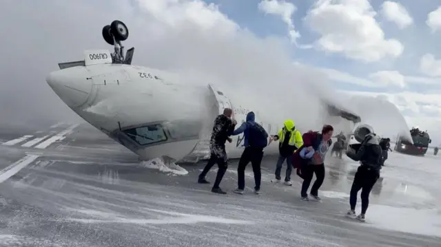 Plane flipped over on its back on snow-covered runway with foam being sprayed at it. People are seen walking away from the aircraft