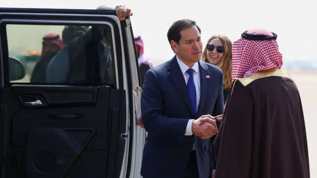 Marco Rubio shakes hands with a man in traditional Saudi Arabian dress, next to a car which has its door still open