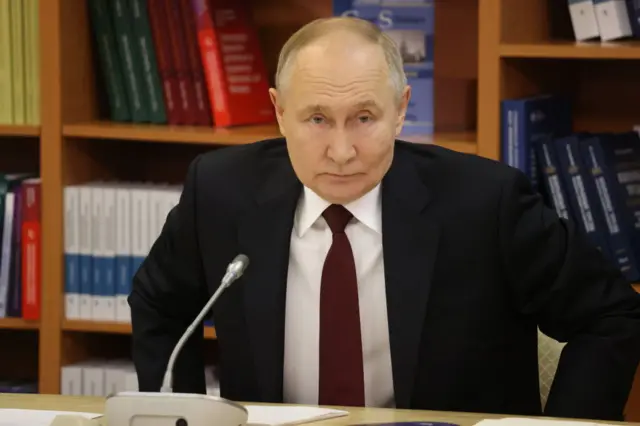 Vladmir Putin sitting in front of a microphone at a desk in front of a bookshelf