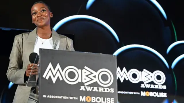 Little Simz stands at a podium with the words Mobo Awards written on it. She is wearing a light green blazer and white t-shirt