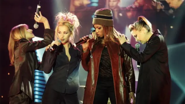 All Saints members Melanie Blatt, Natalie Appleton, Shaznav Lewis and Nicola Appleton sing into microphones on stage. They are all wearing black, with Shaznav Lewis wearing a burgendy leather coat.