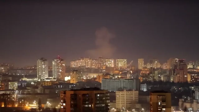 Smoke rising above a city skyline. It's night time so the sky is dark, but there are windows lit up across the city in several high rises. The sky glows a faint orange. In the distance is a large plume of smoke.