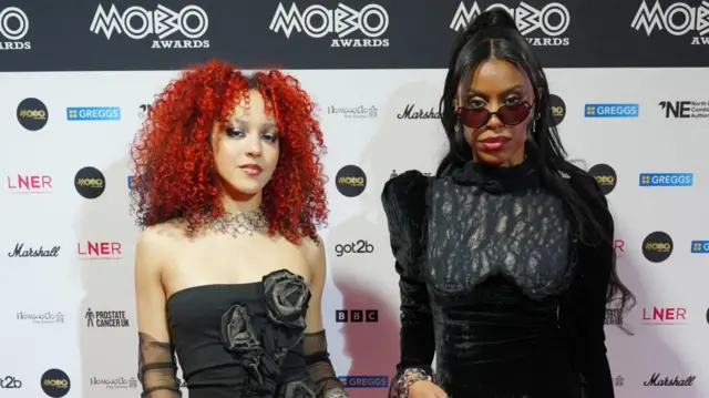 Nova Twins look at the camera on the red carpet. Georgia South, standing on the left, has red hair and is wearing a black strapless dress with flowers on it. Standing next to her is Amy Love, also wearing black with sunglasses.