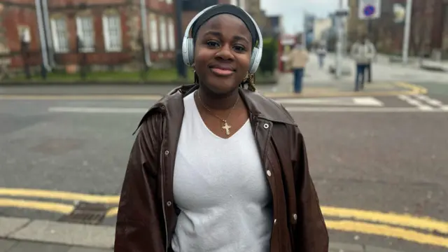 Simi Odemeru smiles for the camera. She is stood on the corner of a street and is wearing white headphones, a grey top, a brown leather jackets, and a golden cross necklace. She has brown hair and brown eyes.