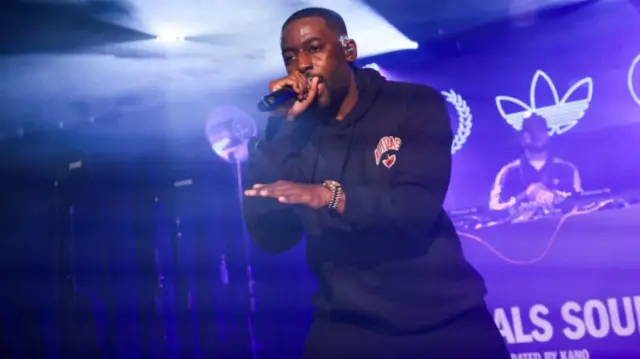 Bashy sings into a microphone on a stage lit by blue and purple lights. He is wearing a black jumper and black jogging bottoms.