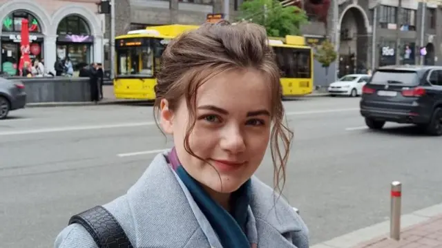 Headshot of a woman standing in front of a street. She has brown hair, that is tied up with two curly bits left out in front of her face. She smiles at the camera