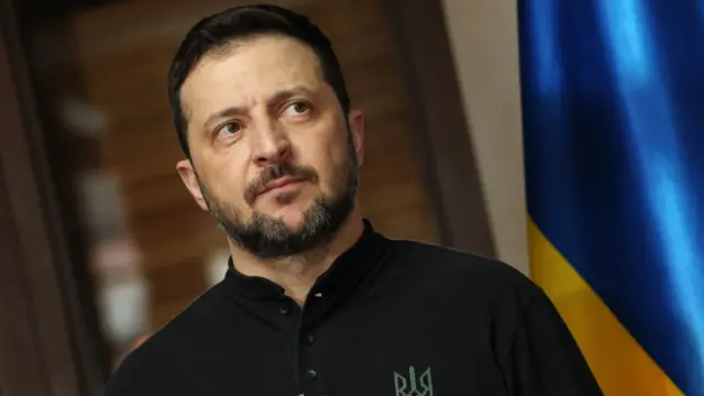 Headshot of Zelensky who looks serious. There's a partial Ukranian flag in view.