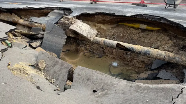 A picture next to the sinkhole where the road has sank and concrete has collapsed into the hole. A pipe now visible where the road has collapsed has a hole in it with water pouring out