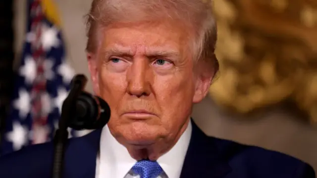 Donald Trump sideyeing, and looking stern and unhappy. He wears a suit and white shirt and blue tie. His face is orange tinged and a microphone is in the foreground near his mouth.