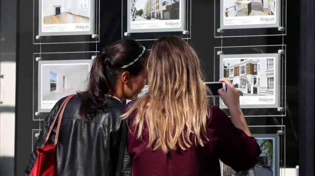 Two women browse adverts for houses for sale in the window of an estate agents