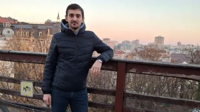 A young man, Oleg Romanyshen, faces the camera as he stands on a bridge overlooking Kyiv, Ukraine.