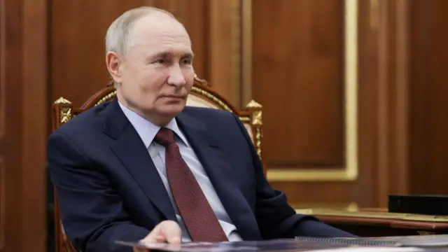 Putin sits back and looks relaxed in a fancy wooden chair. He wears a navy suit, blue shirt and red tie