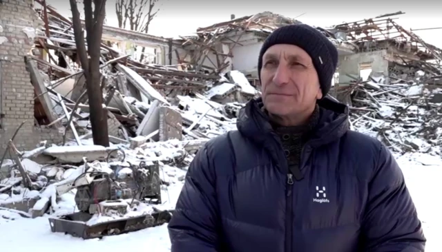 A man wearing a thick black coat and a black beanie stands looking to the side of the camera. Behind him, the ground is covered in snow. There's a ruined building that is covered in rubble, including bricks and many wooden beams