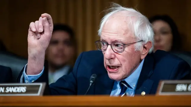 Sanders, sitting at a desk behind a nameplate, raises his right hand as he speaks