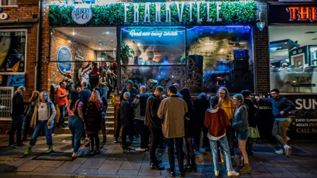 Large groups of people queue up in the dark, outside a lit up music venue