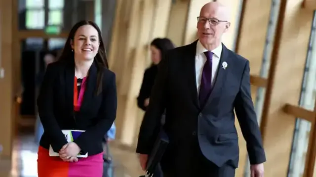 Deputy First Minister Kate Forbes is walking in a Holyrood corridor alongside First Minister John Swinney. Forbes is wearing a two tone skirt with a dark jacket. Swinney is wearing a dark suit and a dark tie, with glasses. Both are smiling.