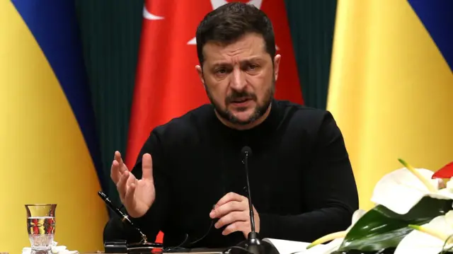 Ukrainian President Volodymyr Zelensky attends an Ankara press conference. He is seated at a table with a water glass and microphone, dressed in black. He has a perplexed expression on his face.