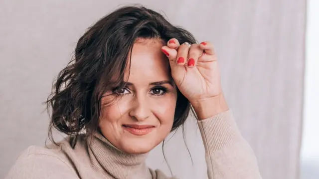 Headshot of a woman smiling at the camera with short brown curly hair. She rests one hand against her forehead, she has red short nails.