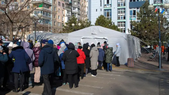 Local people stand in line for construction materials and humanitarian aid near a tent