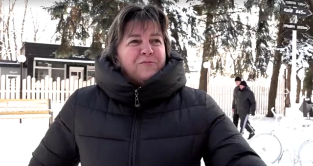 A woman wearing a big black winter coat. There's snow on the ground and trees behind her. Two people walk in the background