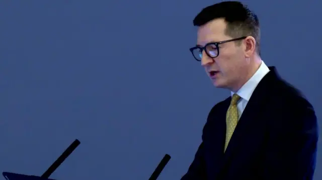 Man wearing black rimmed glasses, a black shirt, a white shirt and a yellow tie.