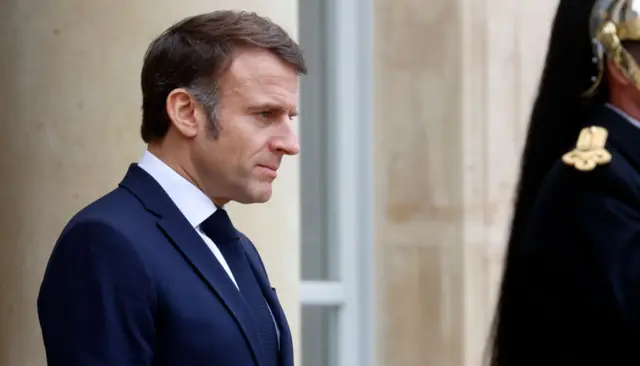 From side on, Macron looks ahead of him with a stern expression. He wears a navy suit and tie, and white shirt.