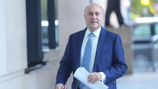 Andrew Griffith in blue suit and tie with papers in his hand walking, blurred background of BBC entrance behind