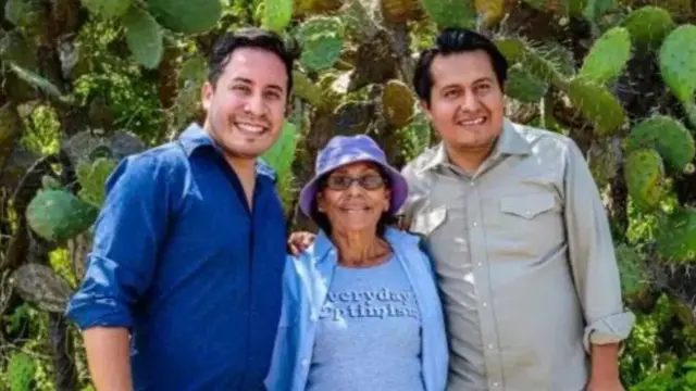 A man in blue shirt has his arms around a woman in light blue shirt and t-shirt with a purple bucket hat. Another man in a light grey shirt stands to the left of the woman, a wall of cacti behind the trio