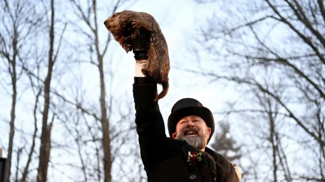A man in black jacket and top hat holding up a groundhog with trees in the background