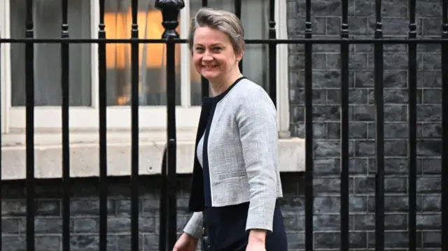 Yvette Cooper on Downing Street in grey blazer and navy skirt, smiling at the camera, standing in front of black railings with a house behind