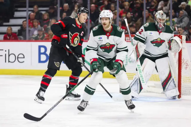 Three ice hockey players - two Minnesota Wild players and one Ottawa Senator player - on the ice with crowds spectating behind them