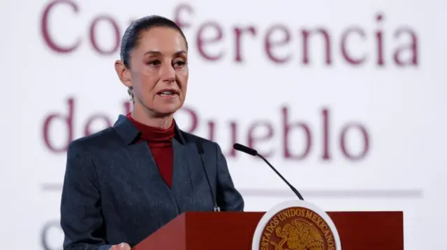Claudia Sheinbaum with her hair pulled back in a chignon deliver a speech stending behind a red-topped lectern while wearing a grey jacket and red turtleneck