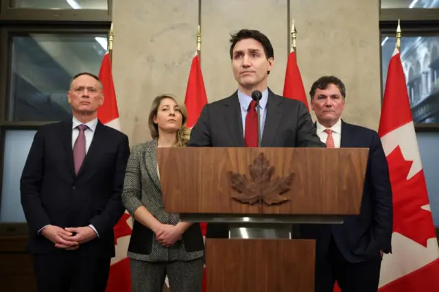 Justin Trudeau stands behind a podium. Other individuals stand behind him with Canadian flags in the background.