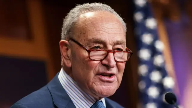 Close up of Chuck Schumer delivering a speech standing. He wears a dark blue-grey jacket, striped white shirt and glasses